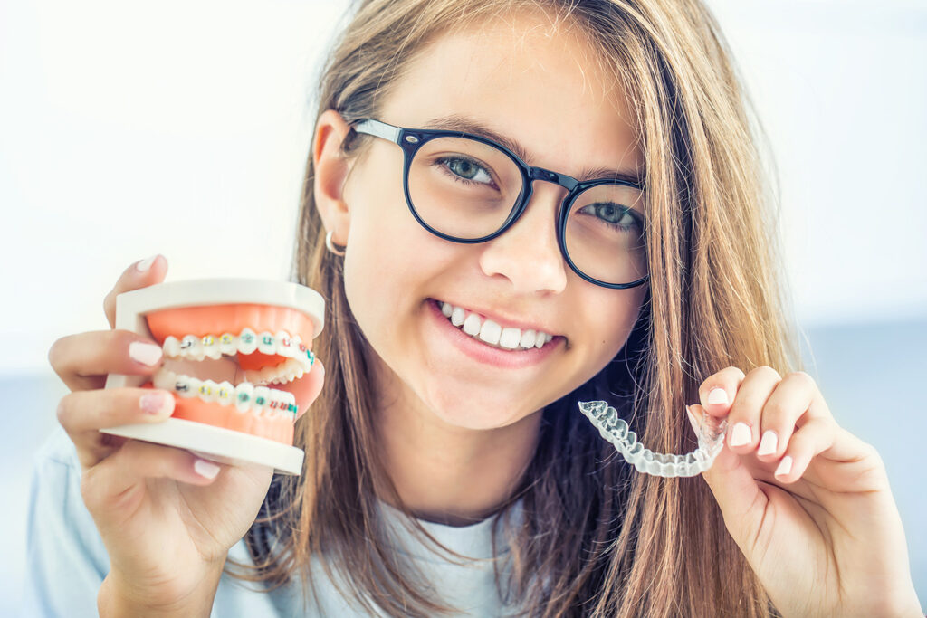 smiling girl holding clear aligners in one hand and mouth model with braces in the other