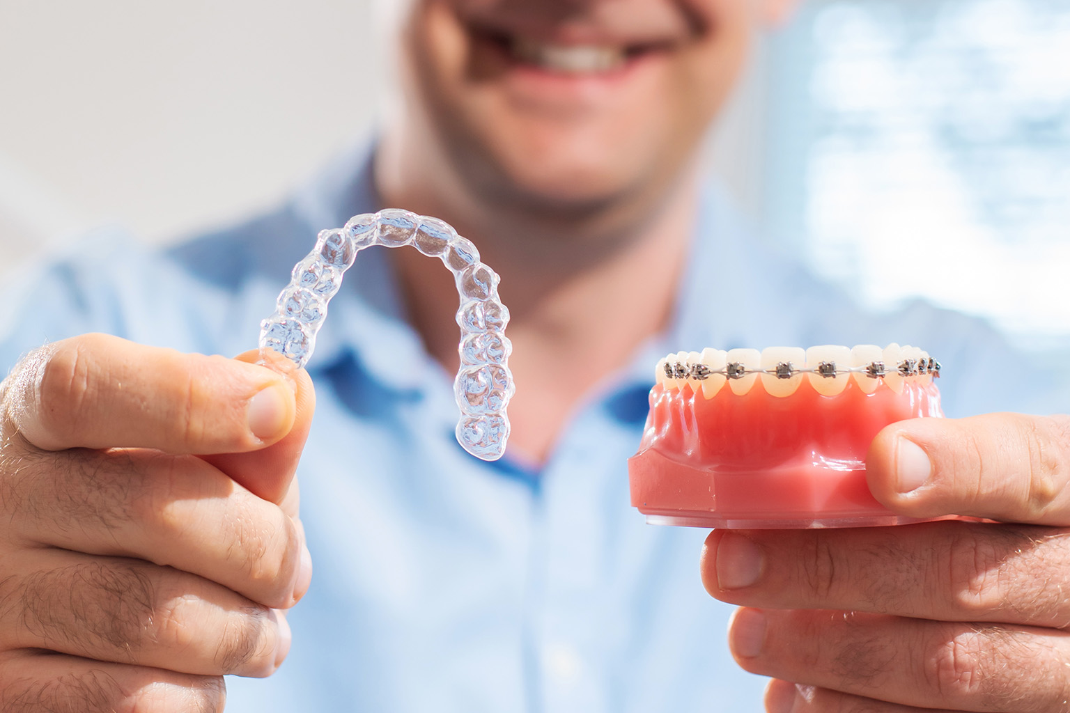 man holding clear aligners in one hand and a model of teeth with braces in the other