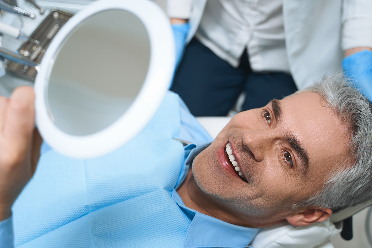 man wearing dental bib smiling into hand mirror