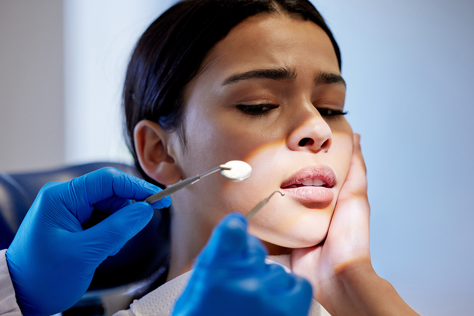 gloved hands holding dental tools towards woman holding her cheek in discomfort