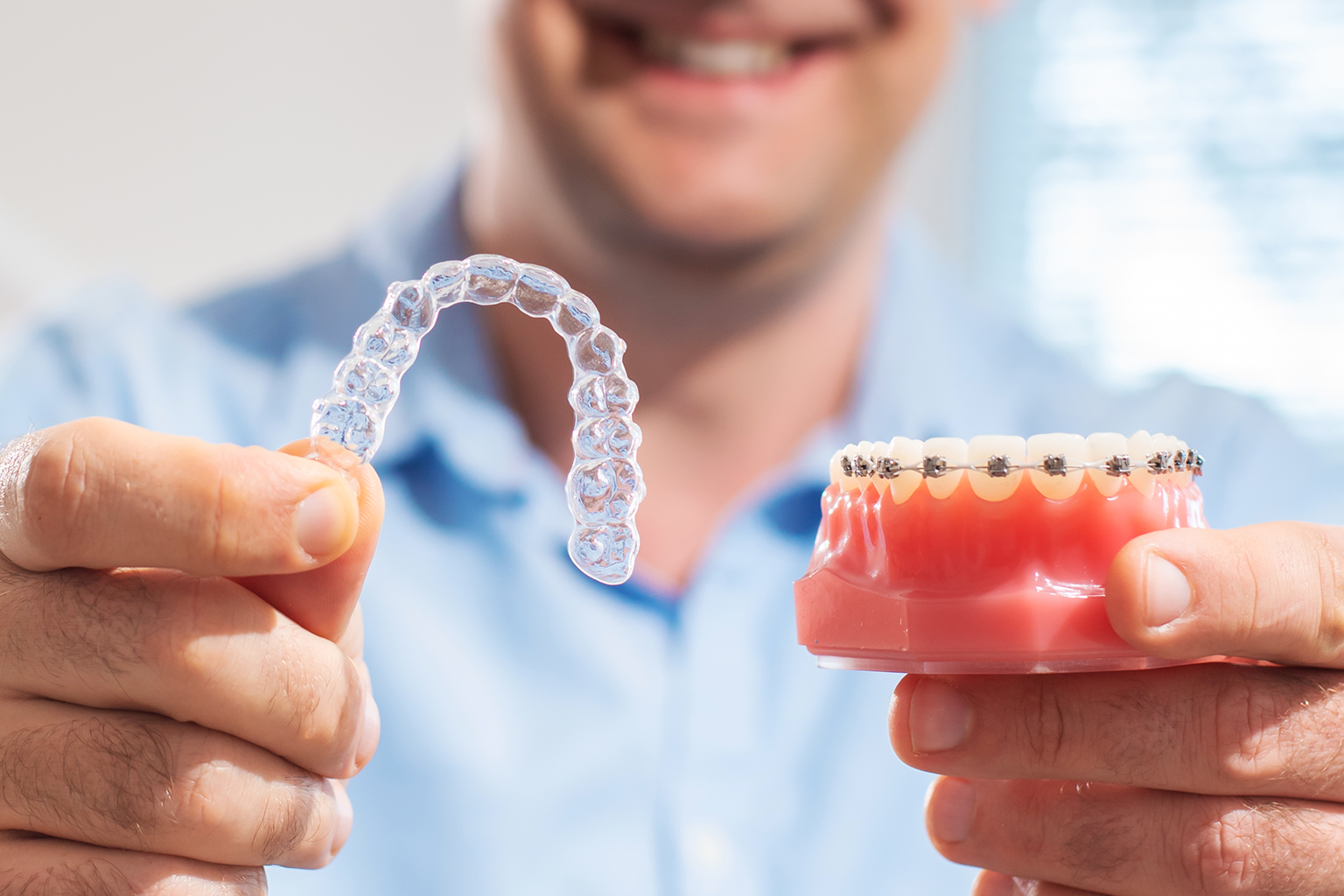 man holding clear aligner in one hand and mouth model with braces in the other