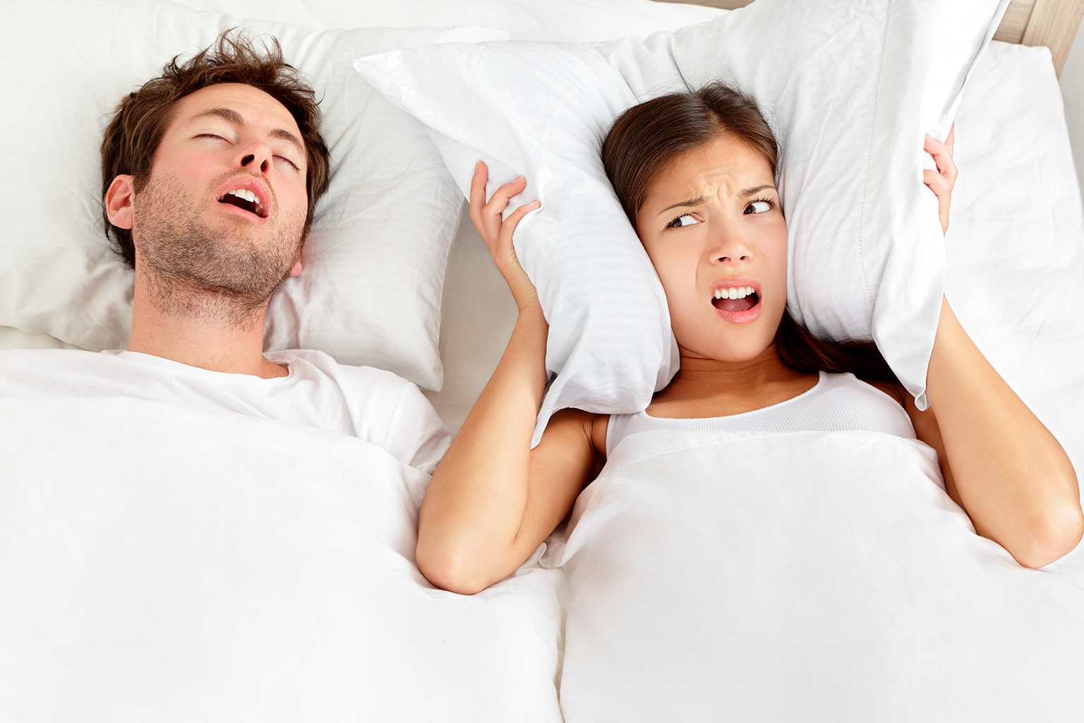 woman covering her ears with a pillow beside sleeping man with his mouth open