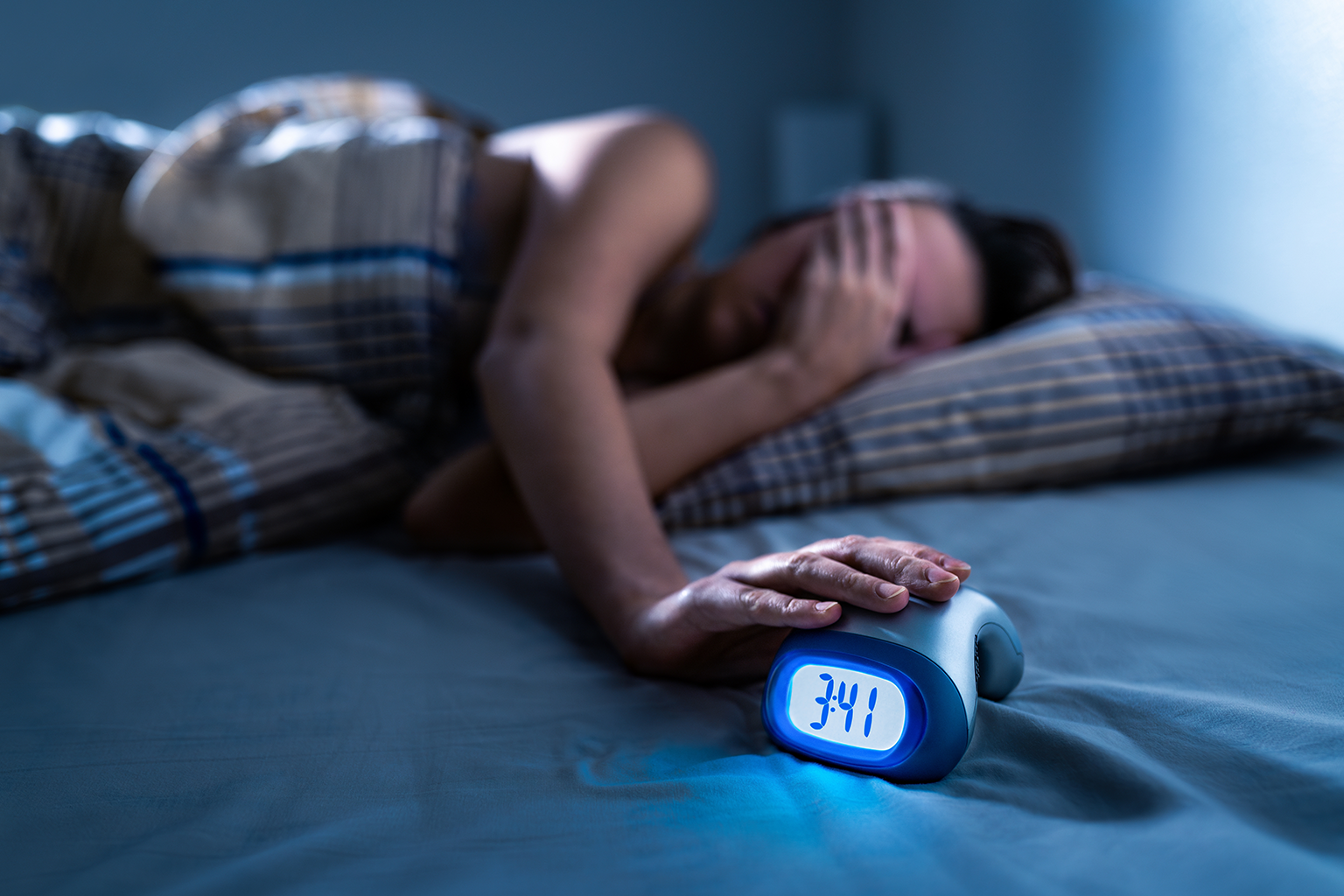 person in bed covering their face with their hand on a digital clock