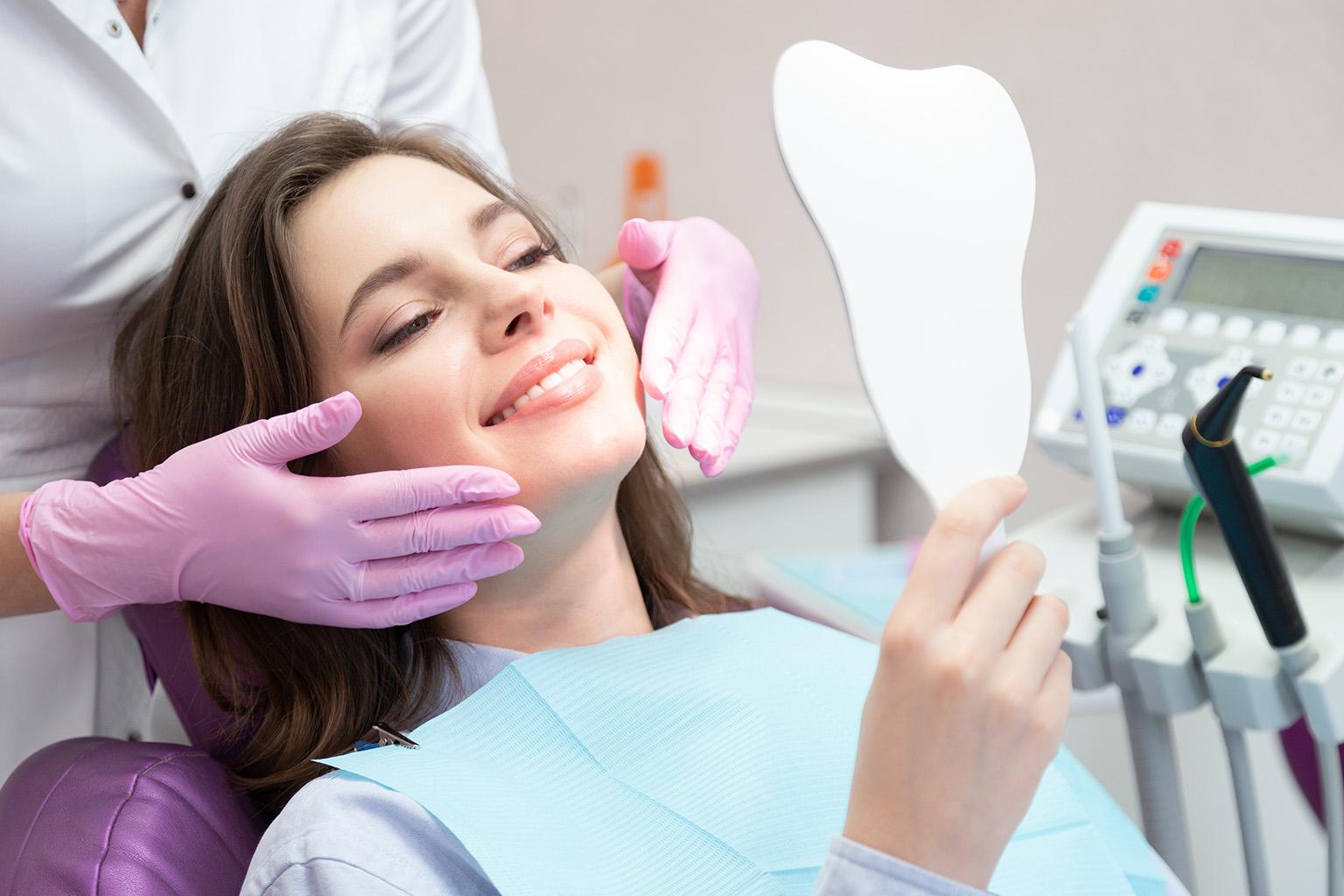 woman in dental chair smiling into hand mirror while gloved hands frame her face