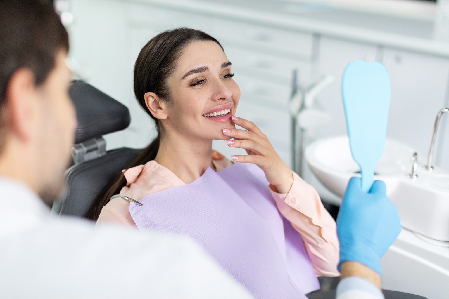 woman touching chin and smiling into mirror held by gloved hand of male dentist