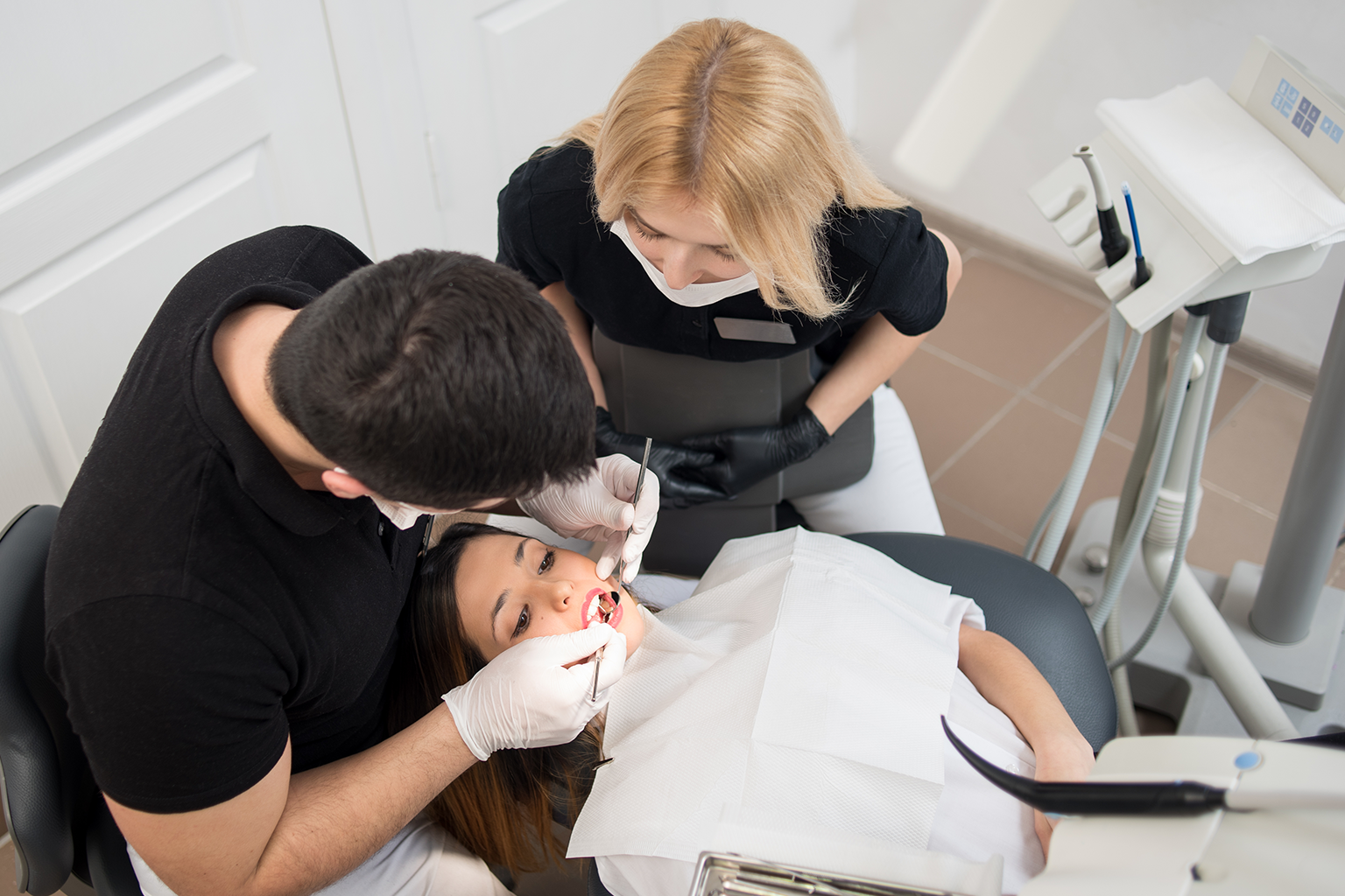 Dentist using dental tools in mouth of patient in dental chair while assistant watches.