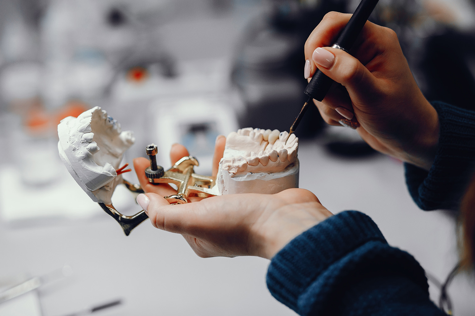 Hands using dental tools to shape tooth replacements on jaw model.