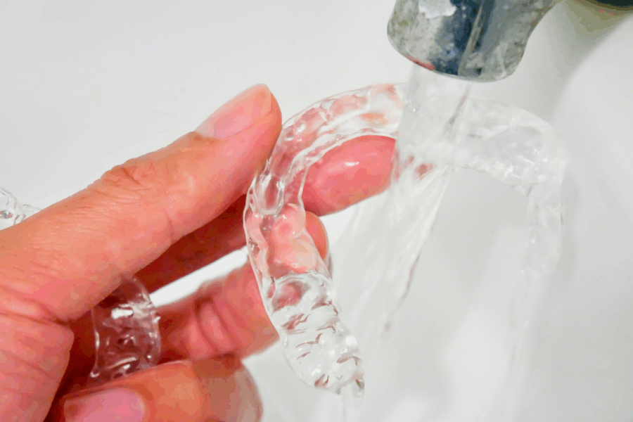 Hand holding clear aligner under water faucet.